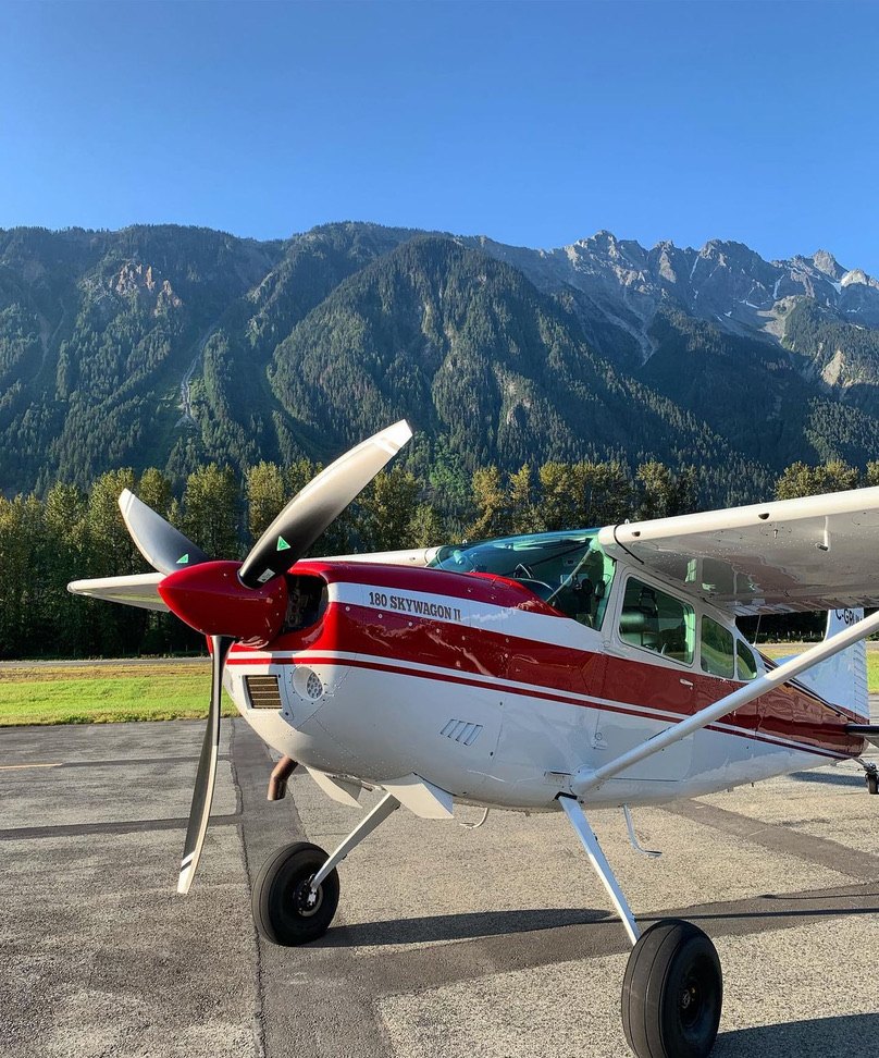 Red and white Cessna 180 Skywagon II with a three-blade MT propeller on the ramp in front of mountains