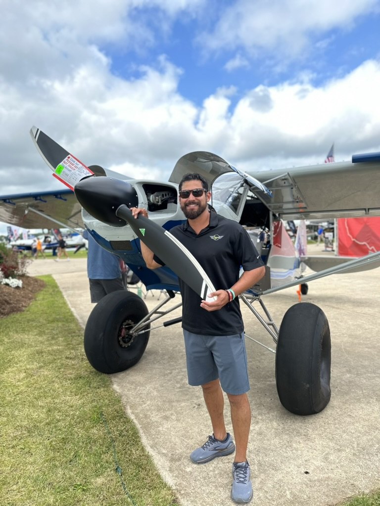 Smiling man in sunglasses holding a black MT propeller blade in front of a silver and blue bush plane