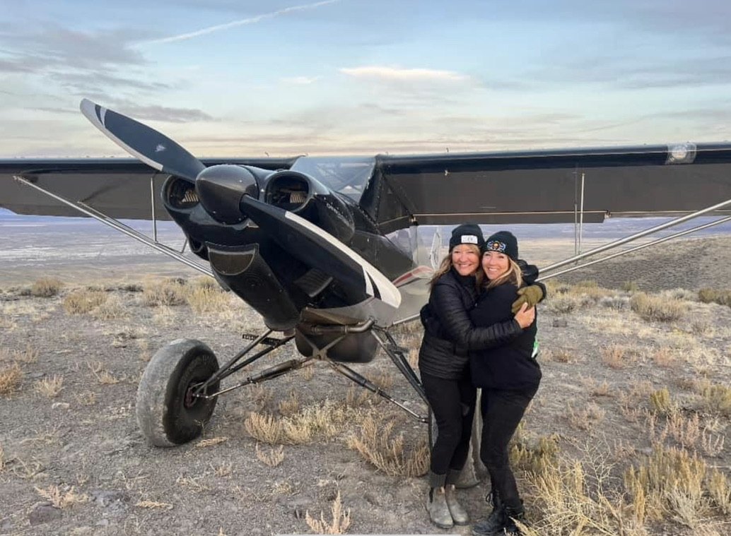 Two women hugging in front of a black bush plane with a black MT propeller in the desert
