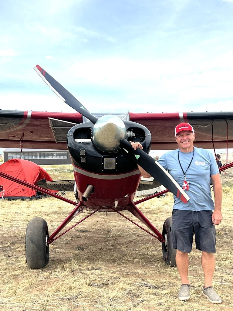 Smiling man with a red cap standing beside a red and black Aviat Husky with a black MT propeller