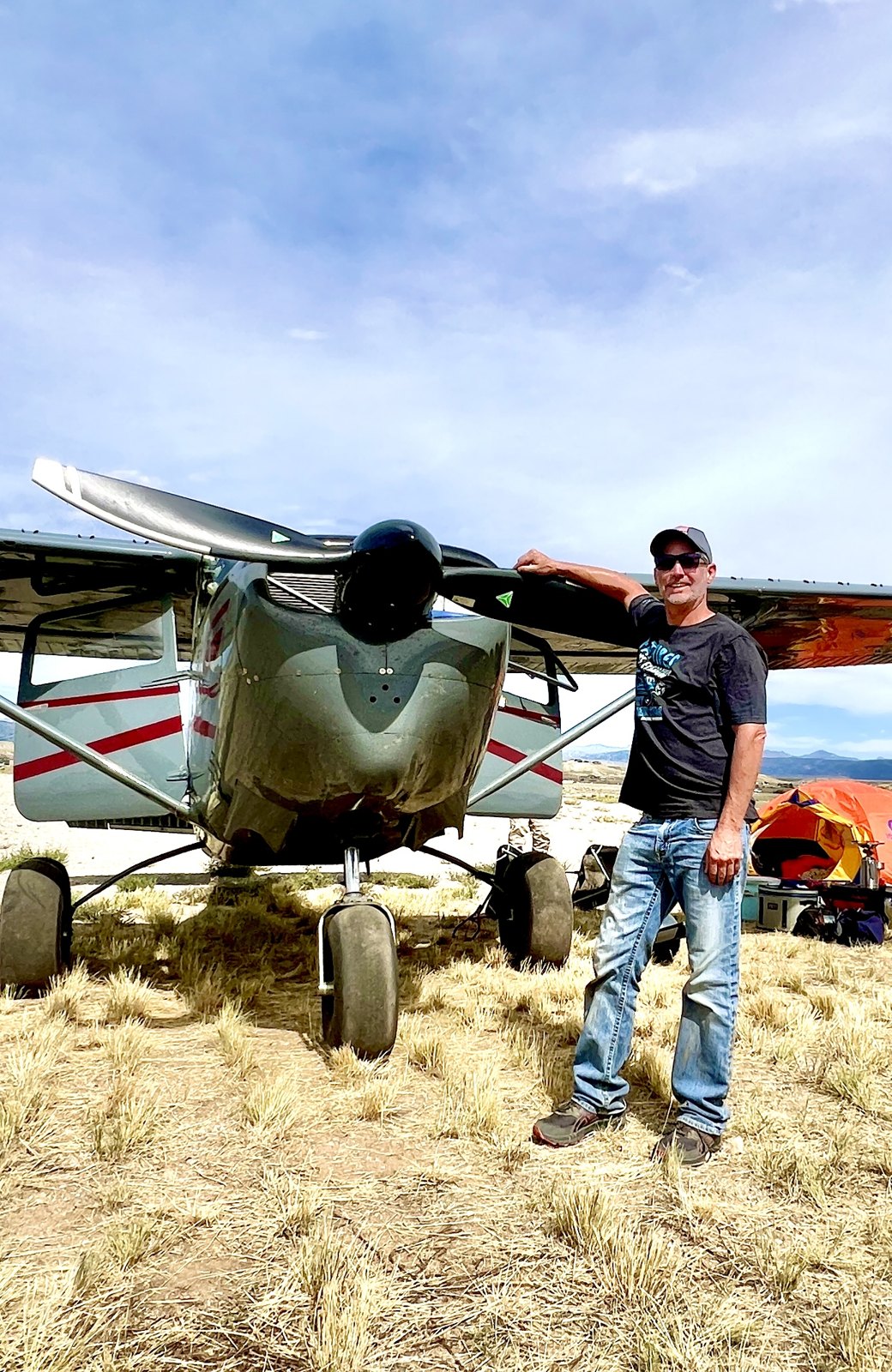 Man in sunglasses leaning on the black three-blade MT propeller of a gray Cessna on a dry grass strip