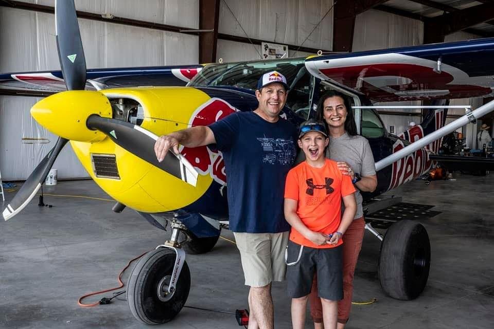 Family of three smiling in front of a yellow, blue and red Cessna with a black MT propeller in a hangar