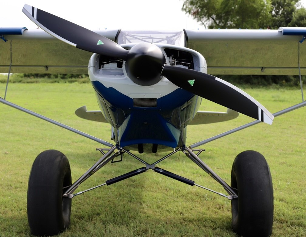Front view of a silver and blue bush plane on grass with a black two-blade MT propeller