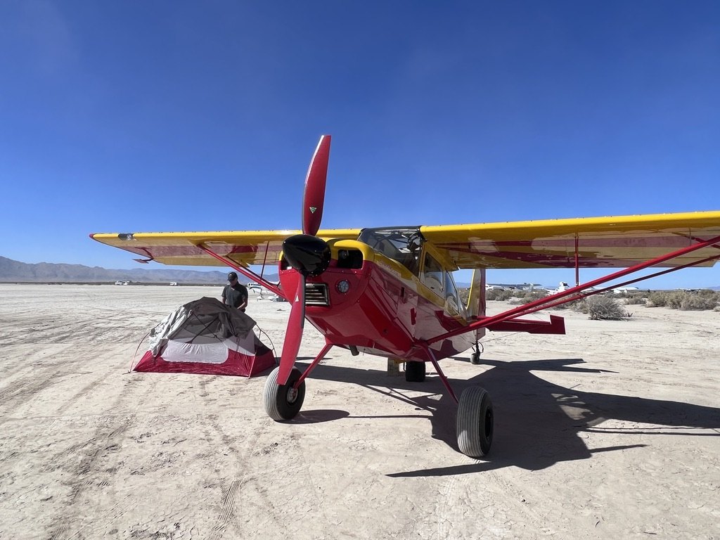 Red and yellow bush plane with a black MT propeller on a dry lakebed with a tent