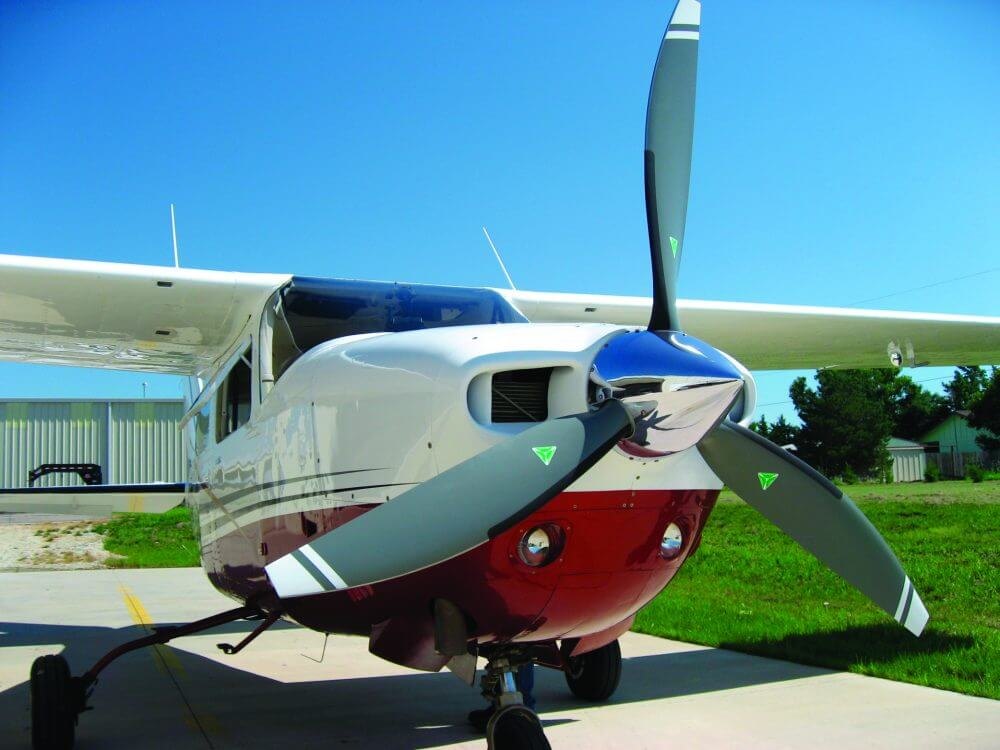 Maroon and white Cessna 210 with a gray three-blade MT propeller in front of a hangar