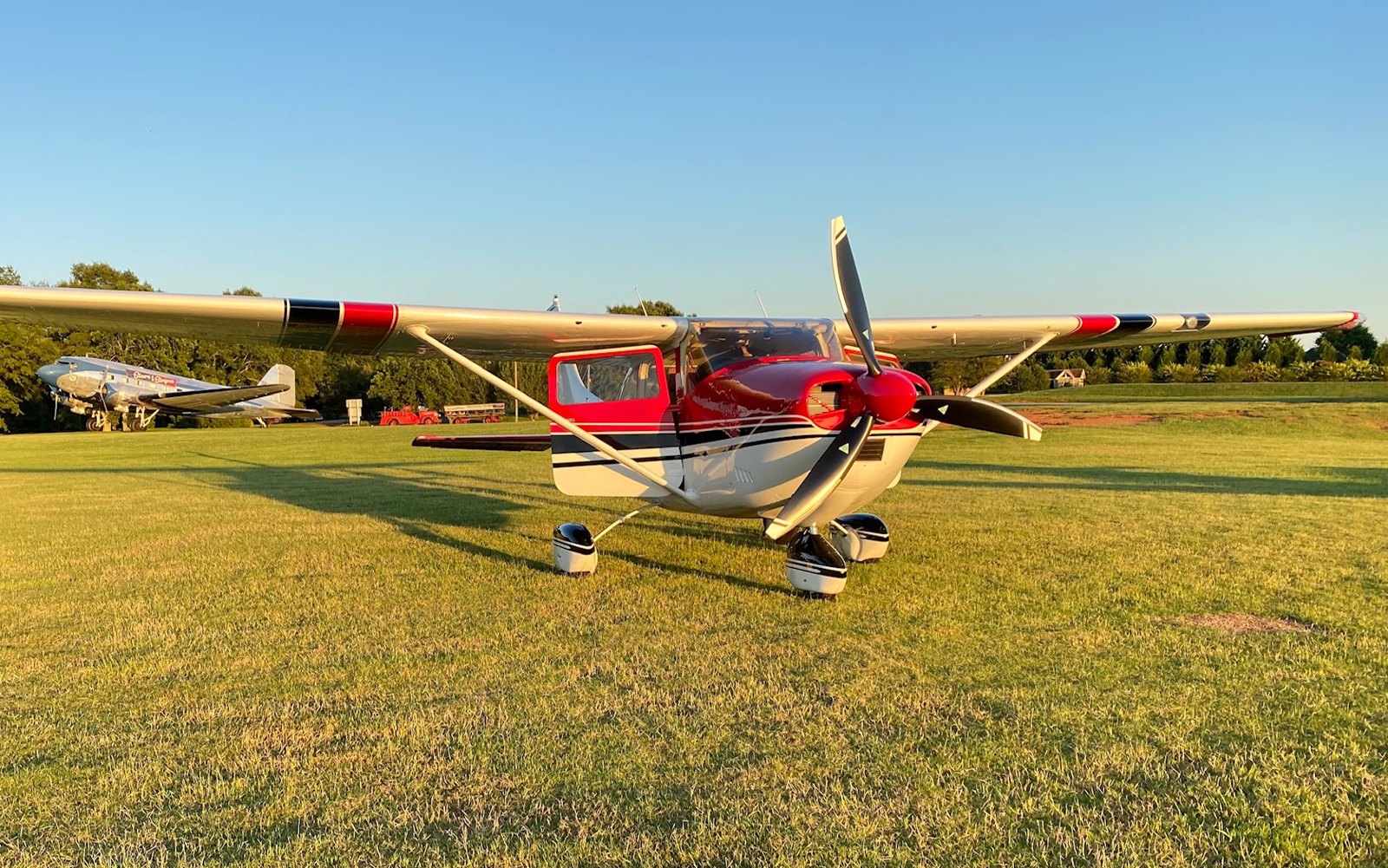 Red and white Cessna with a three-blade MT propeller on a grass strip at sunset