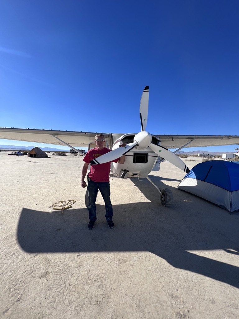 Man in a red shirt standing in front of a white Cessna with a white three-blade MT propeller on a lakebed