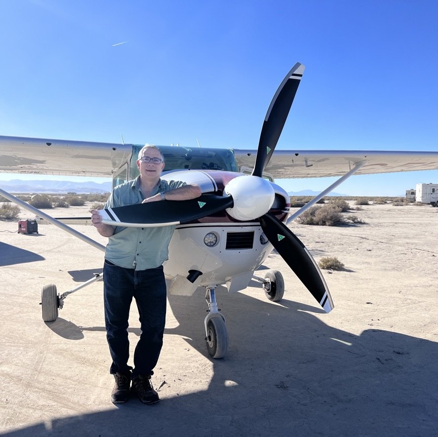 Man in glasses leaning on the black three-blade MT propeller of a Cessna 182 on a dry lakebed