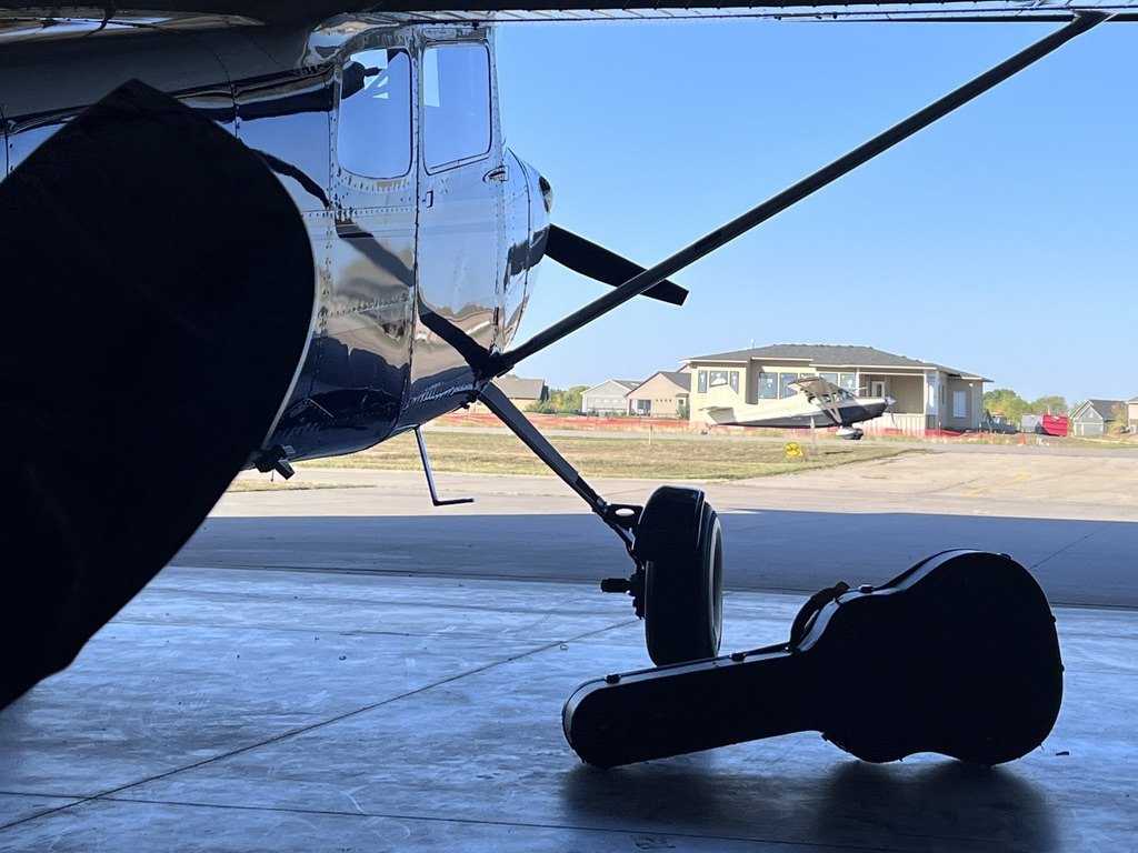 View from inside a hangar of a polished Cessna and a guitar case with a plane parked outside
