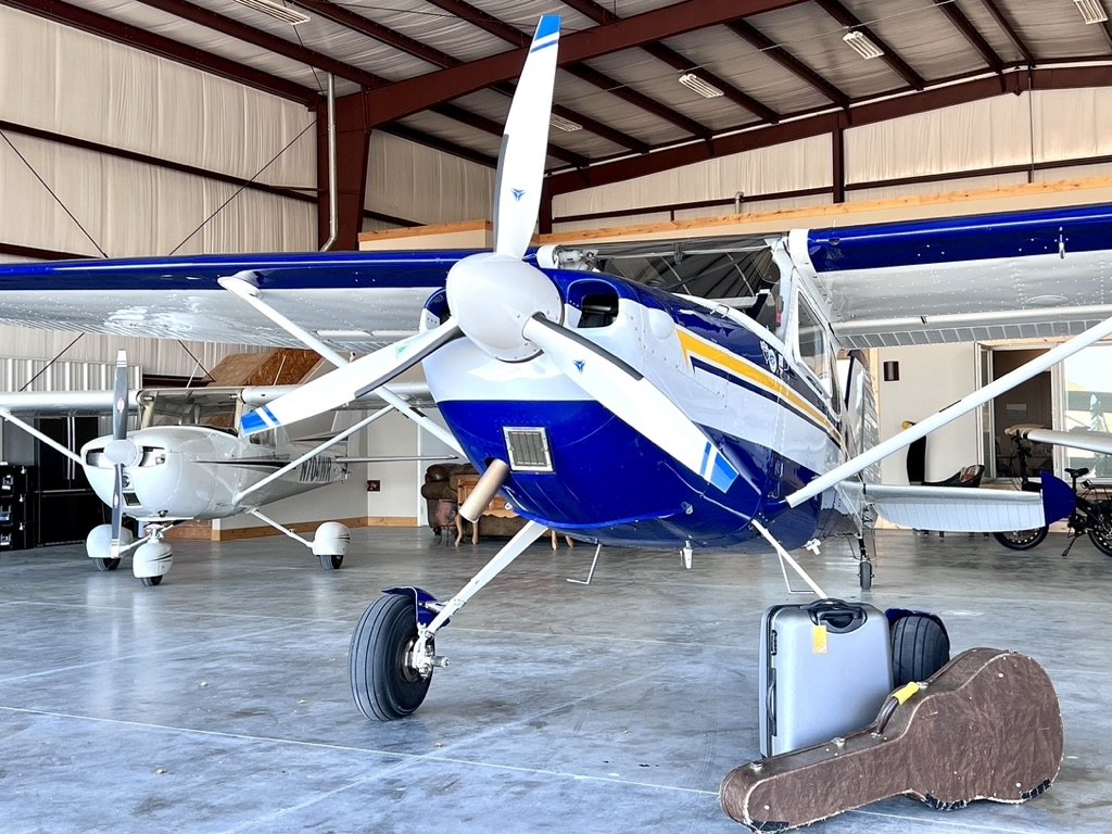 Blue and white Cessna with a white three-blade MT propeller inside a hangar