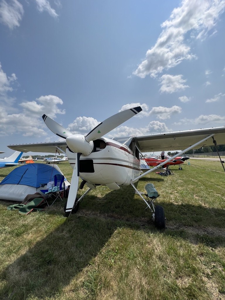 White Cessna with a white three-blade MT propeller parked in the grass beside a tent