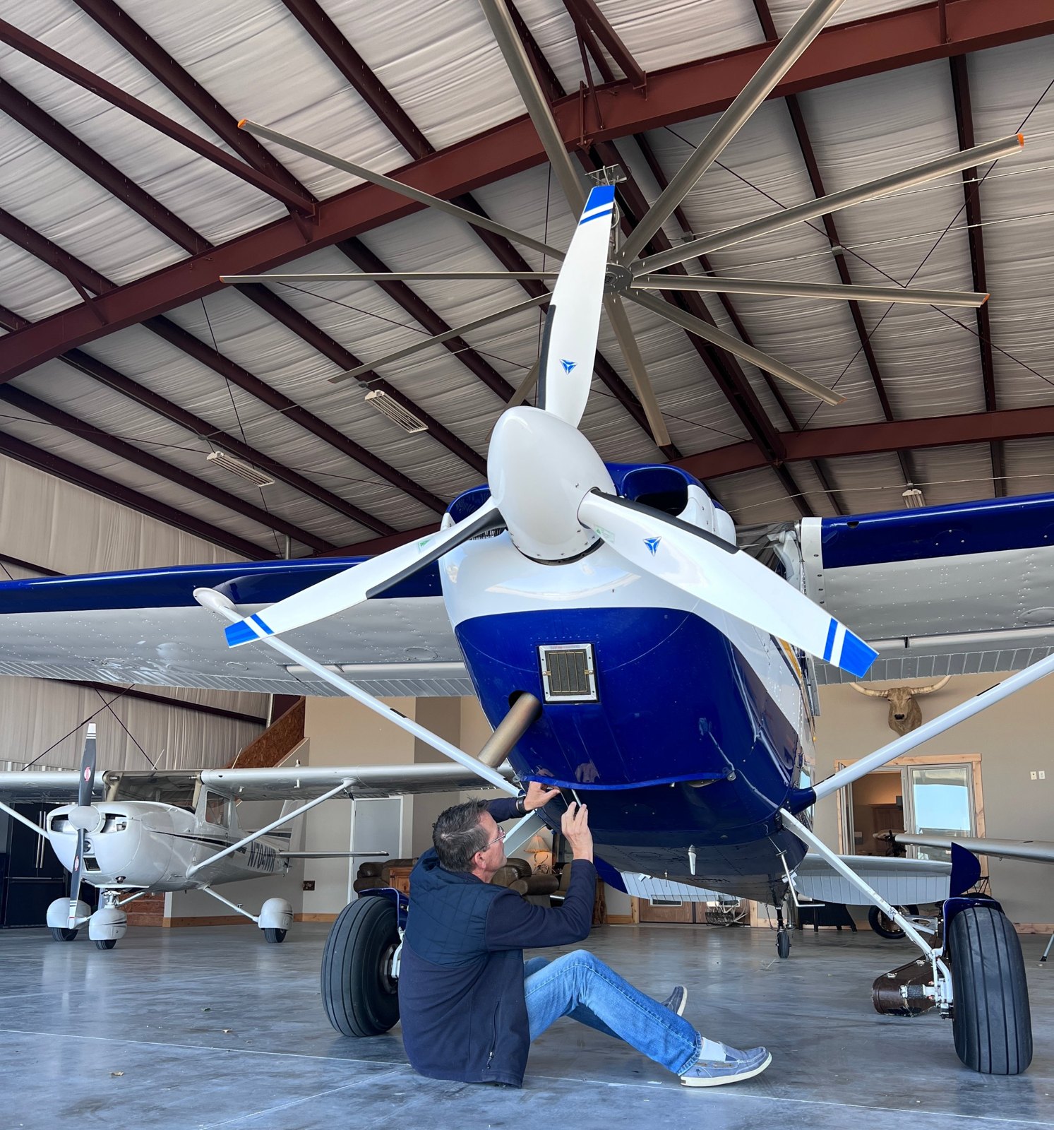Man sitting on the hangar floor checking the underside of a blue and white Cessna with a white three-blade MT propeller