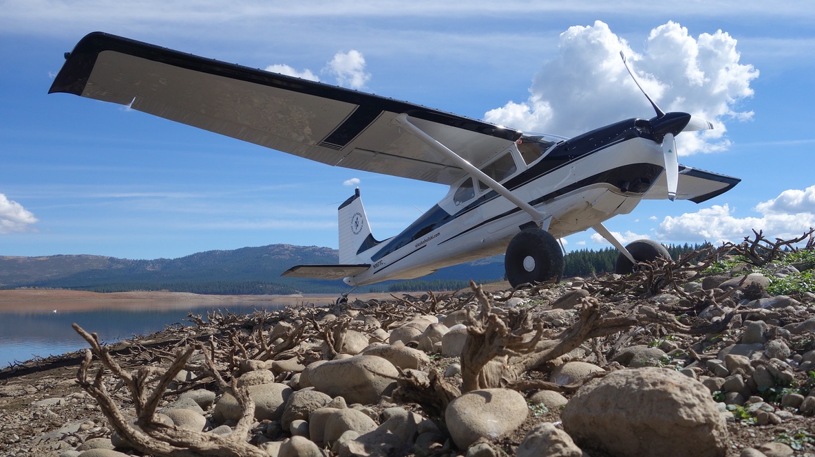 Cessna 180 on a gravel lakeshore beside a reservoir, with a black MT three-blade propeller, under blue sky and clouds
