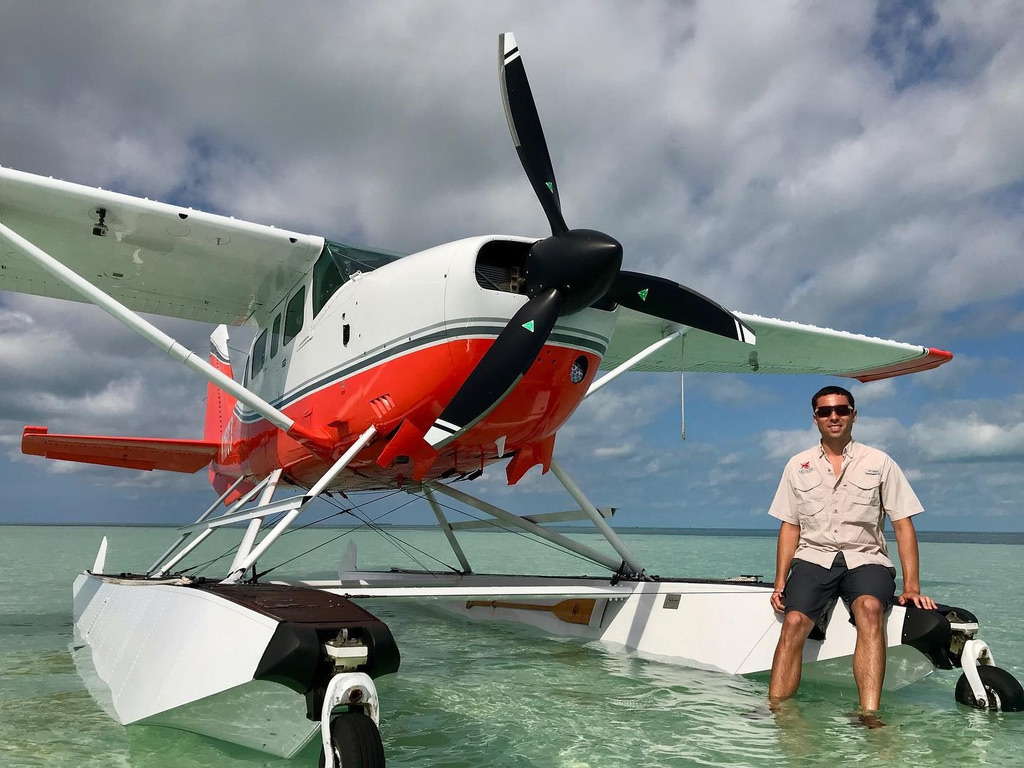 Red and white Cessna 206 floatplane with a black three-blade MT propeller on clear shallow water, with a man sitting on the float