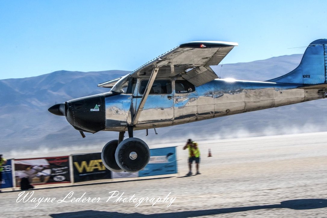 Polished chrome Cessna 180 landing on a dry lakebed with a black MT propeller, mountains behind