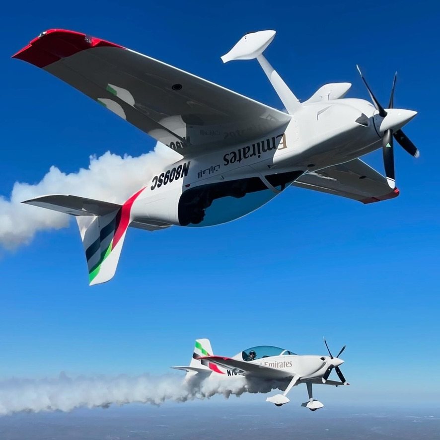 White Emirates aircraft flying inverted above a second Emirates aircraft, with white smoke trails and a black MT propeller
