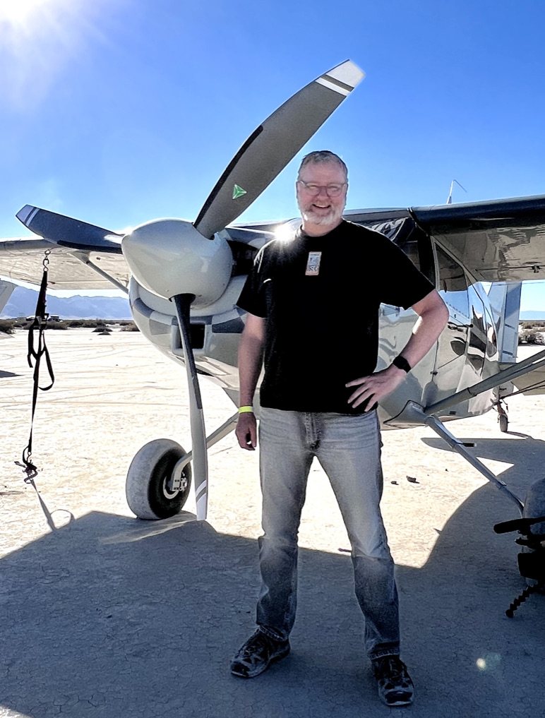 Smiling man standing in front of a Cessna 185 with a gray two-blade MT propeller on a dry lakebed