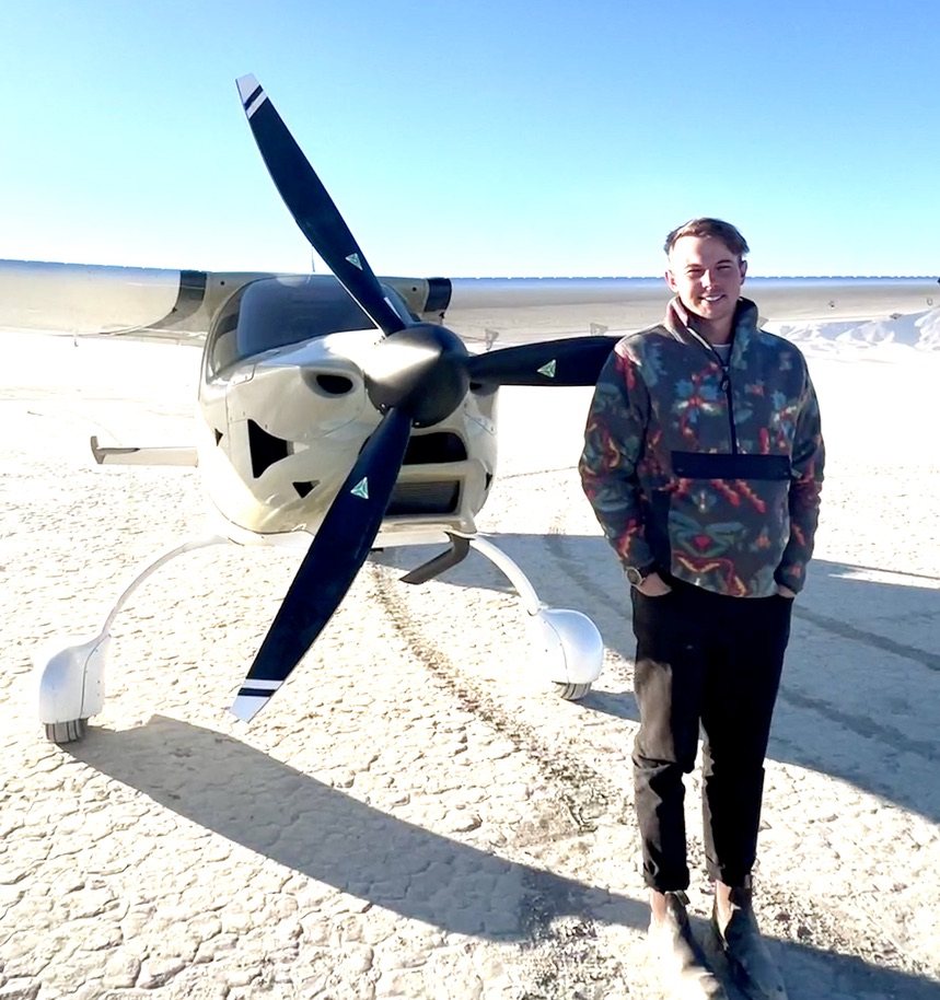 Smiling man in a patterned fleece standing beside a white aircraft with a black three-blade MT propeller on a dry lakebed