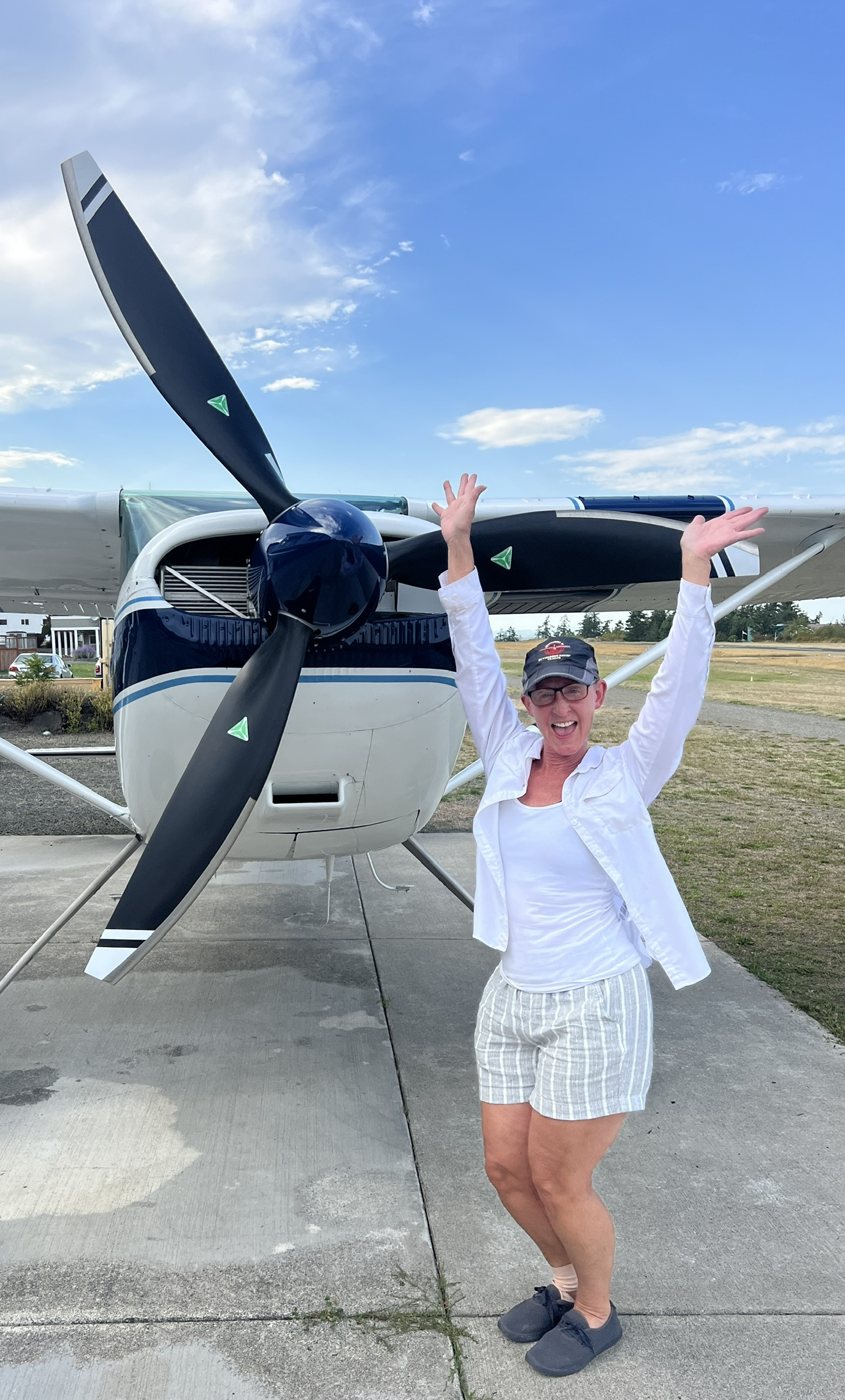 Woman with her arms raised in front of a blue and white Cessna with a black three-blade MT propeller