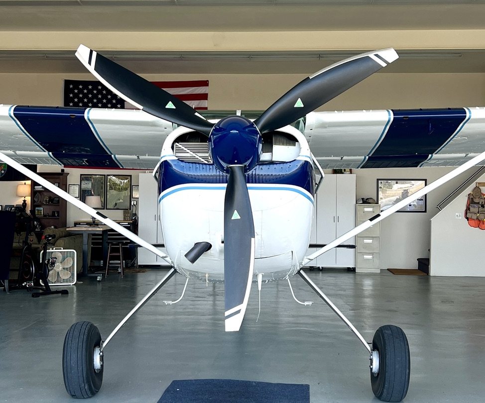 Front view of a blue and white Cessna in a hangar with a black three-blade MT propeller and an American flag behind