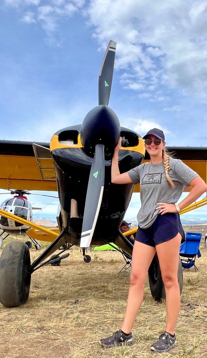 Smiling woman in a gray Bushwheels shirt standing in front of a black and yellow bush plane with a black MT propeller