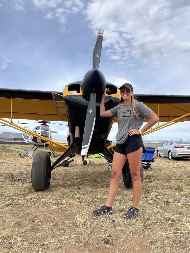 Woman in a gray shirt and cap standing in front of a black and yellow bush plane with a black MT propeller