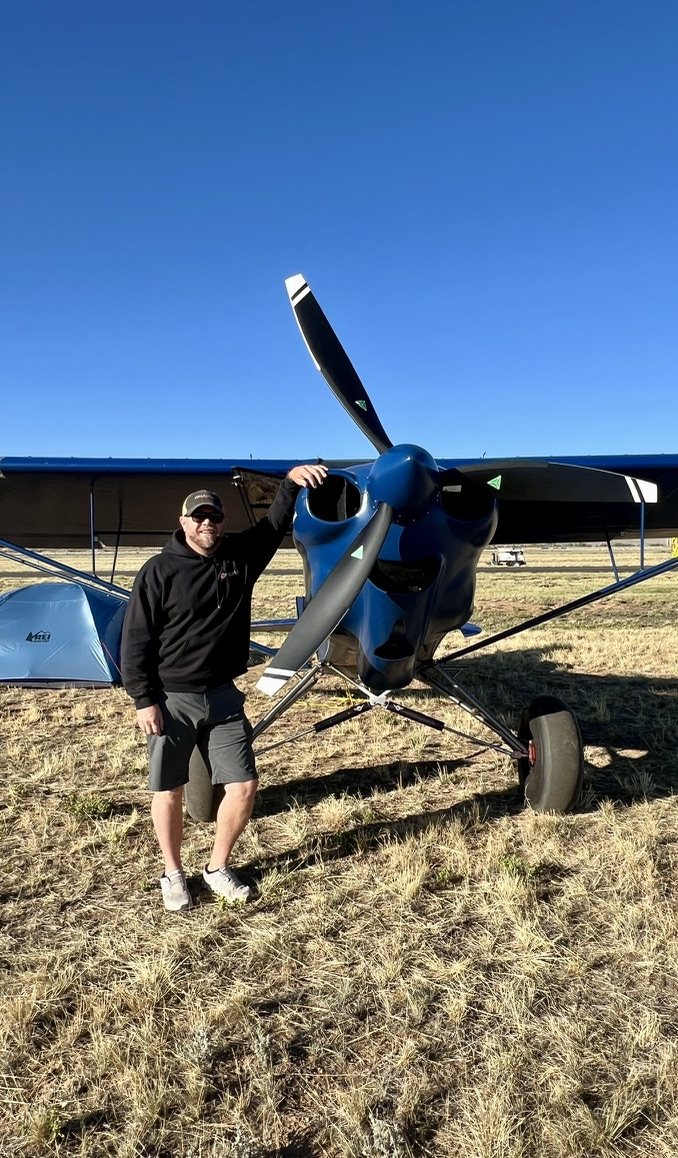 Smiling man in a black hoodie standing beside a blue bush plane with a black three-blade MT propeller, with a tent beside it