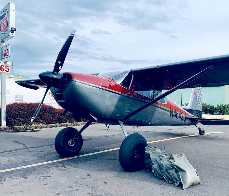 Gray and red Cessna 180 with a black two-blade MT propeller parked beside a Phillips 66 sign