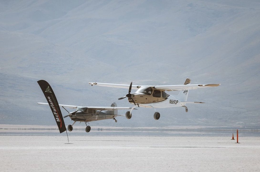 White Cirrus and a Cessna flying low side by side over a dry lakebed with a McFarlane flag