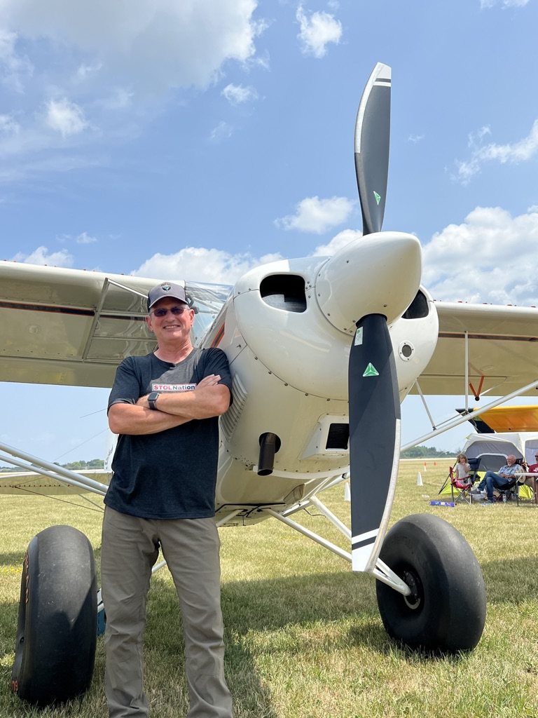 Smiling man with arms crossed beside a white bush plane with a black two-blade MT propeller